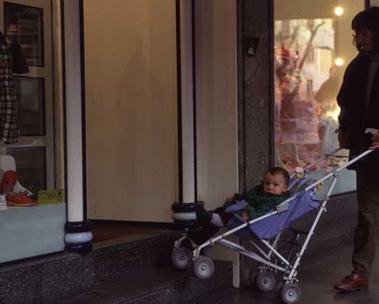 Adulto con bimbo in passeggino che cerca di superare alcuni gradini per entrare in un negozio
