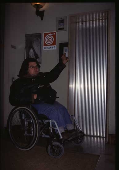 Persona in carrozzina che cerca di raggiungere i pulsanti di chiamata di un'ascensore